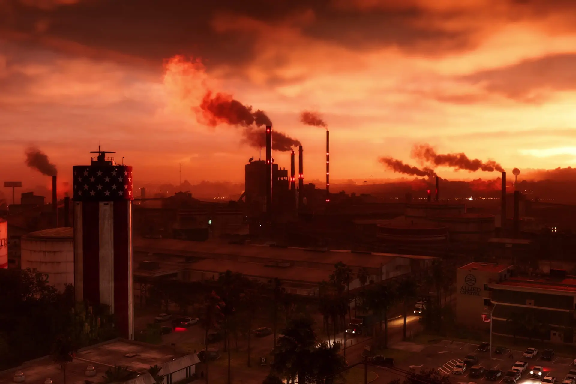 Industrial city at sunset under red-orange skies, smokestacks releasing plumes, a tall American-flag-striped tower and parked cars.